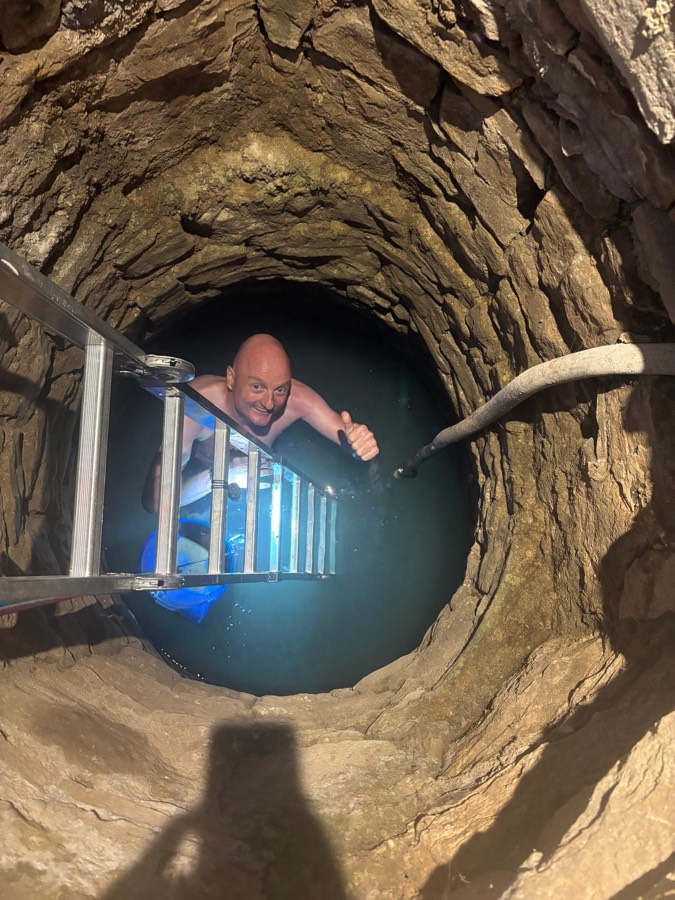 Bram met opgestoken duim in het water van een oude, diepe stenen put met koud bergwater